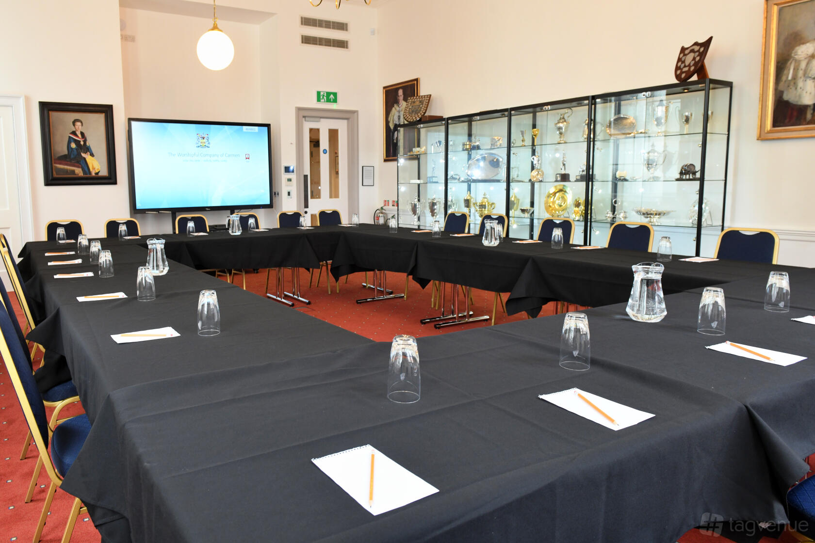 An event space with a U-shaped table layout, black linens, glass cabinets displaying silverware, and framed art at Worshipful Company of Carmen.