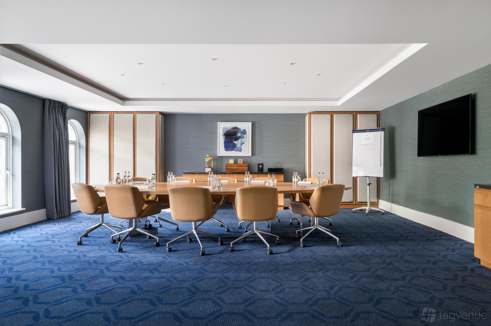 An event suite with a long wooden table, leather chairs, floor-to-ceiling windows, and a large screen at Crowne Plaza Gerrards Cross.