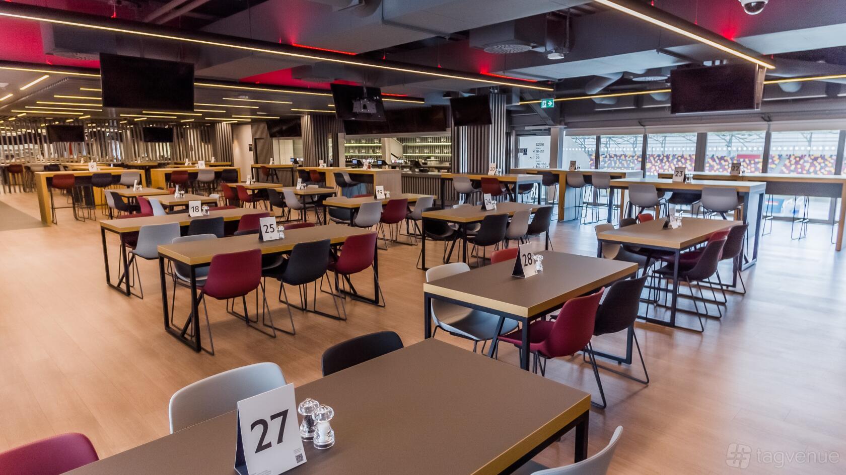 A stadium lounge with numbered tables, mix of red and gray chairs, large windows overlooking the pitch at Brentford Football Club.