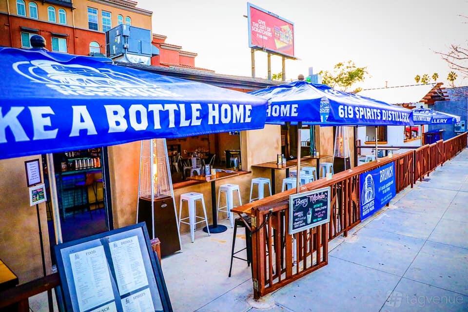 An outdoor restaurant patio with blue branded umbrellas, high stools, and wooden fencing at 619 Spirits North Park.
