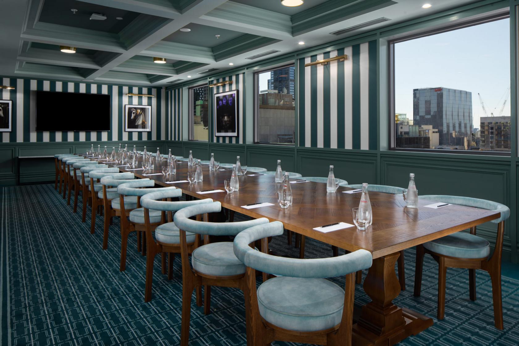 A hotel dining room with a long wooden table, upholstered chairs, striped walls, and large windows at Pullman Melbourne City Centre.