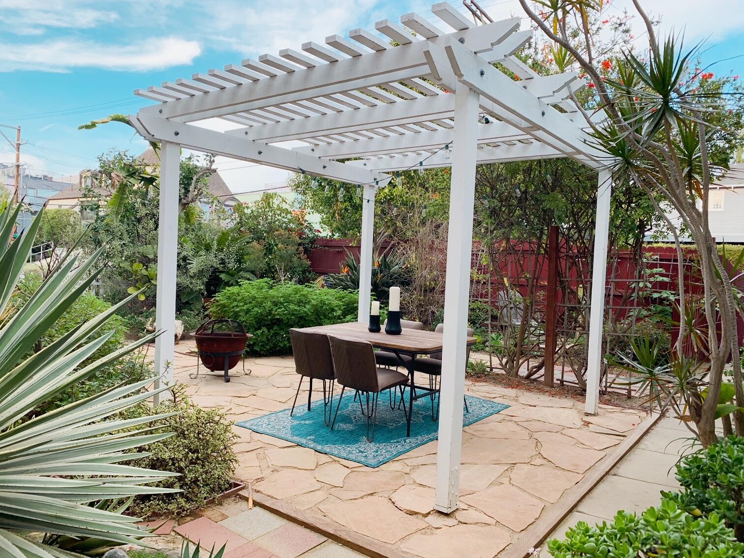 A patio with a white pergola, dining table and chairs on a blue rug surrounded by lush greenery at Beautiful Venice Canal House