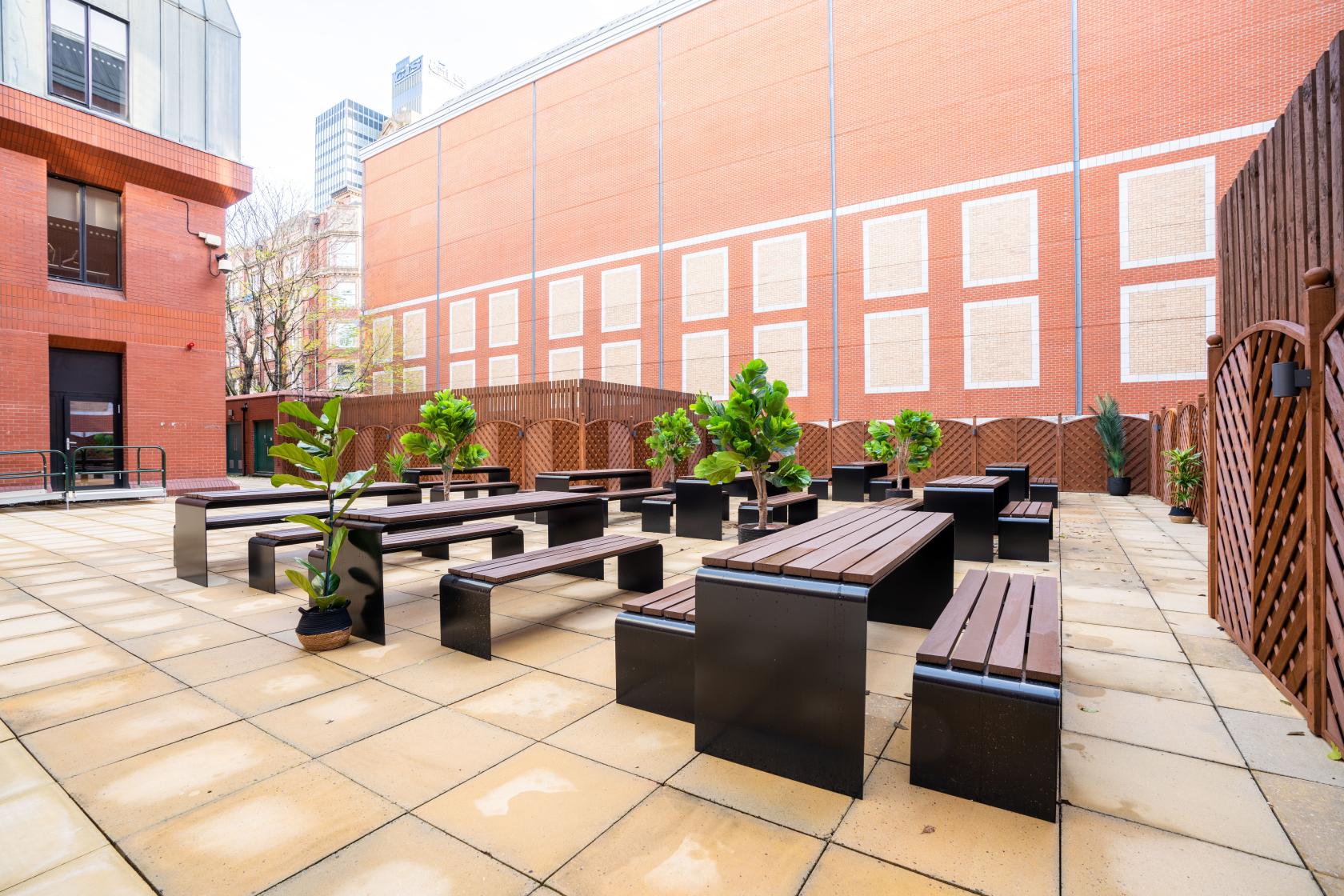An outdoor meeting centre courtyard with wooden benches, tables, potted plants, and brick walls at Orega Balloon Street.