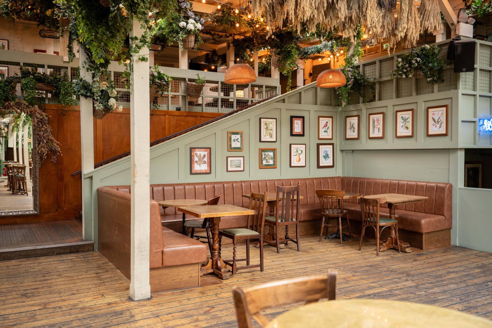 A pub with a large corner banquette, wooden tables, framed botanical prints, and hanging greenery at The Prince West Brompton.
