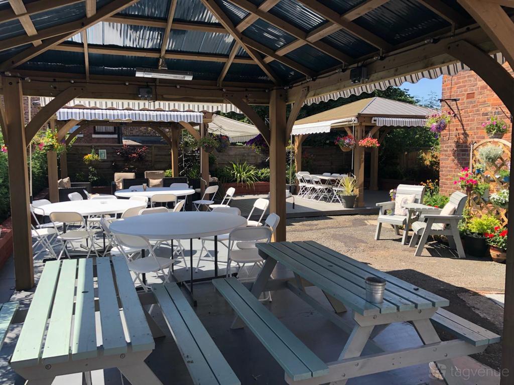An outdoor terrace with covered gazebos, picnic benches, round tables, and potted plants at The Urban Courtyard Manchester.