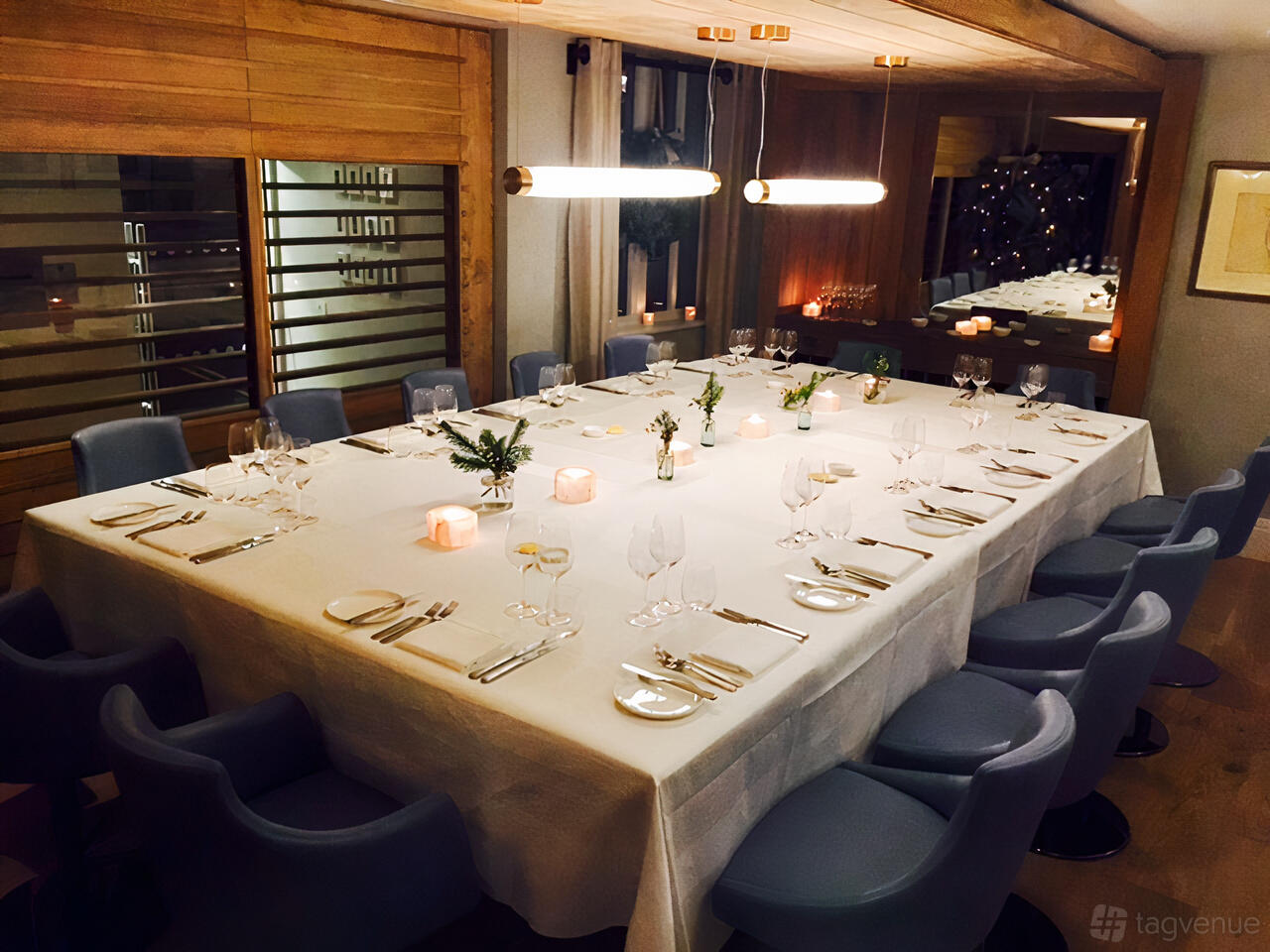 A dining room with a large square table set with white linens, candles, and grey chairs at Elystan Street Restaurant.