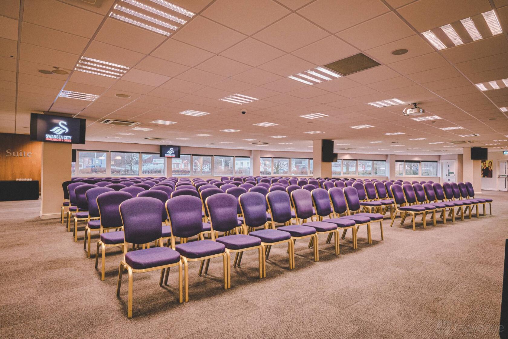 An auditorium with rows of purple chairs, ceiling-mounted projectors, and large windows at Swansea.com Stadium.