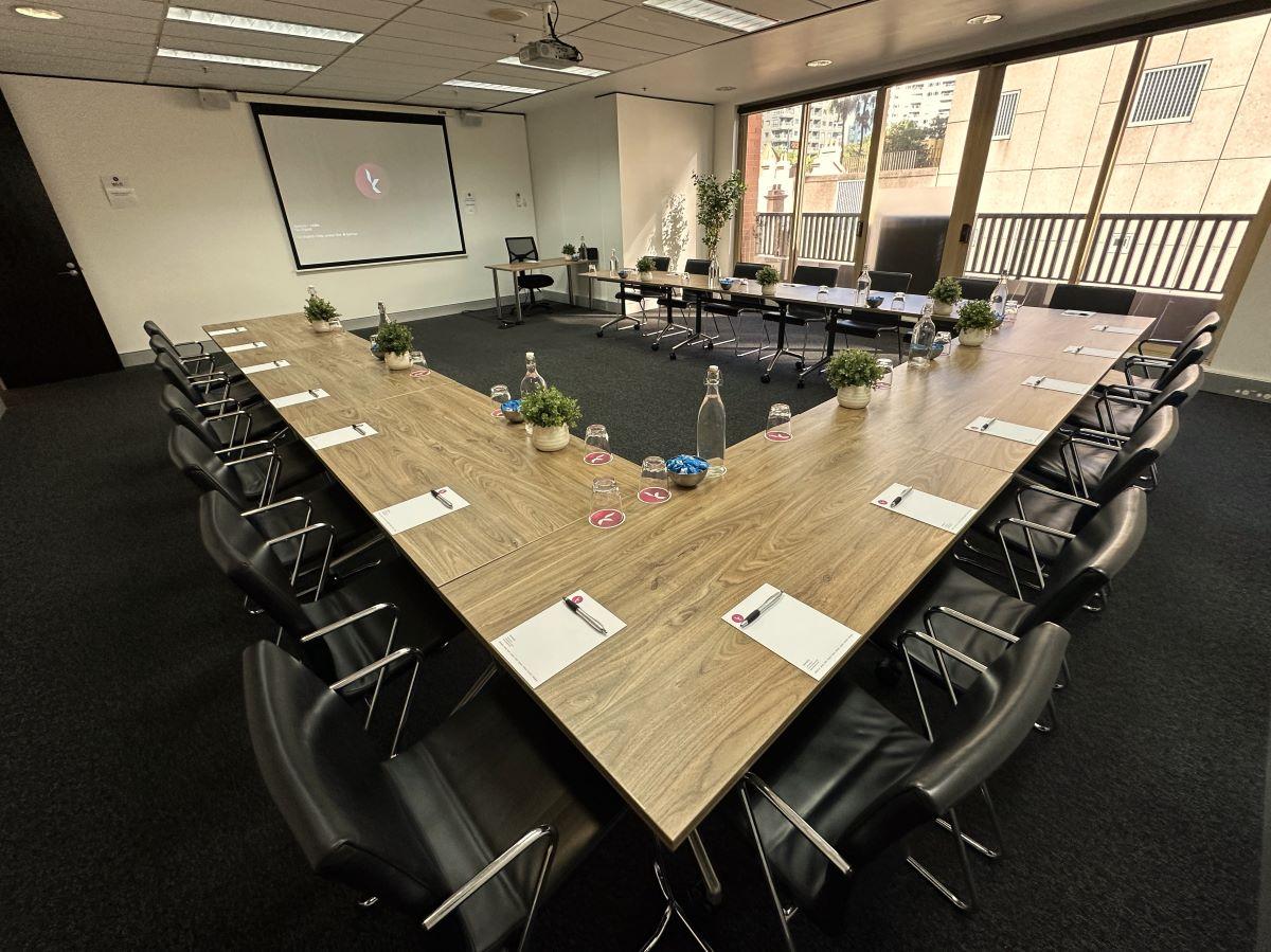 A conference centre room with U-shaped wood tables, black chairs, potted plants, and natural lighting at Karstens Sydney.