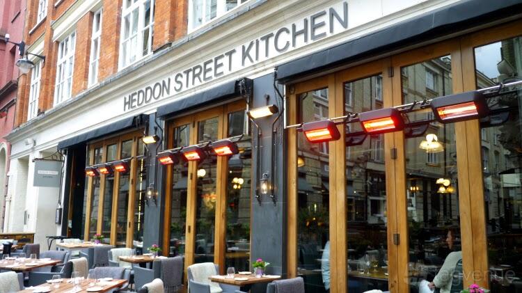 A restaurant with outdoor seating, wooden tables, cushioned chairs, and overhead heaters at Heddon Street Kitchen.