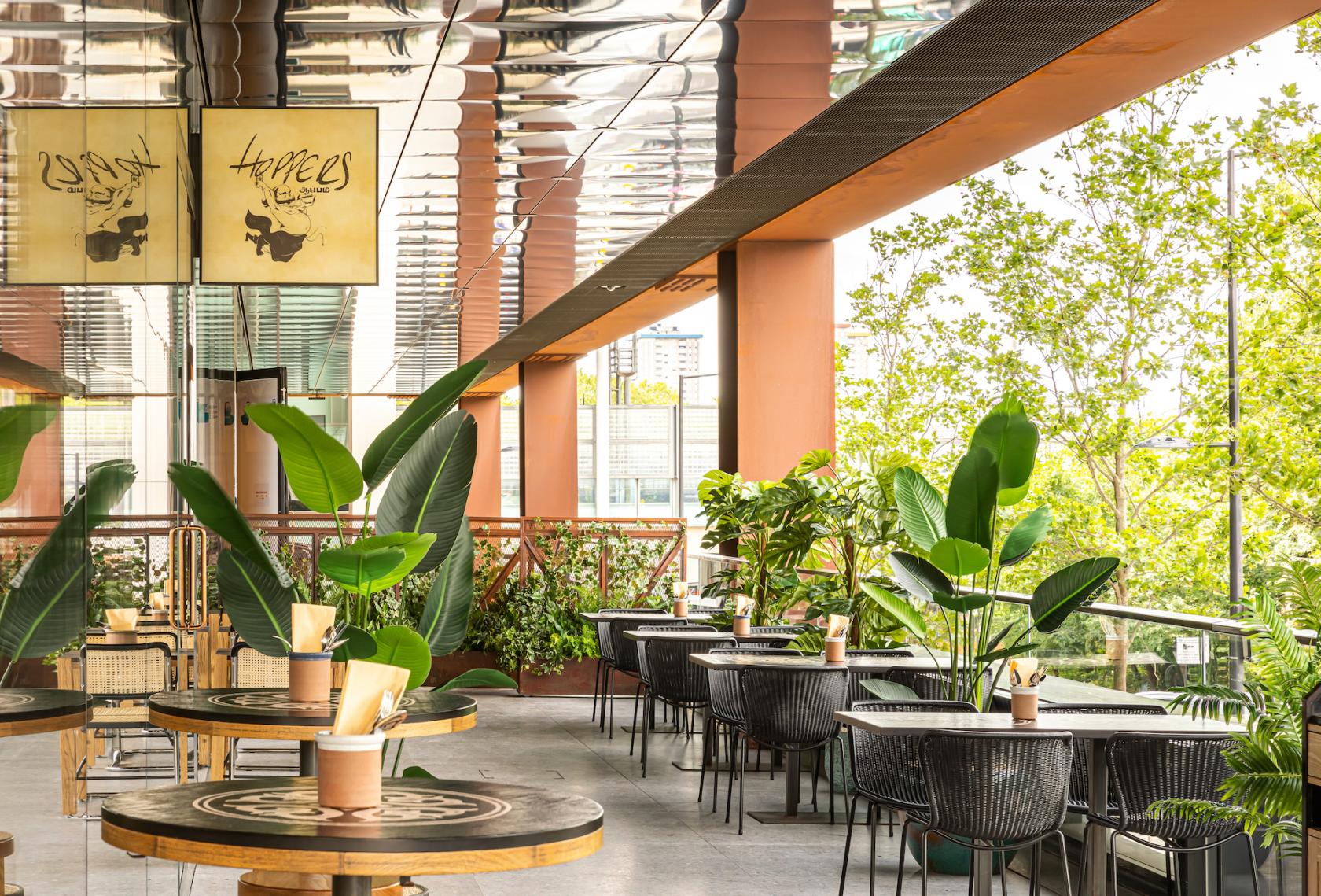 A restaurant terrace with round wooden tables, black wicker chairs, and tropical plants at Hoppers King's Cross.