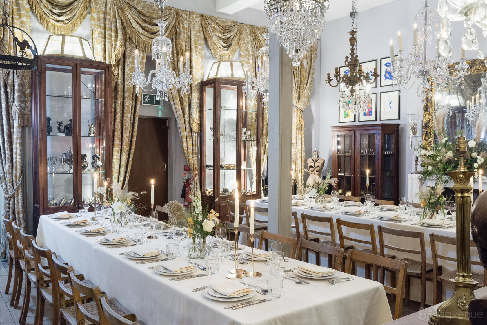 A dining room with long tables, elegant chandeliers, gold drapes, wooden chairs, and display cabinets at Brunswick House.