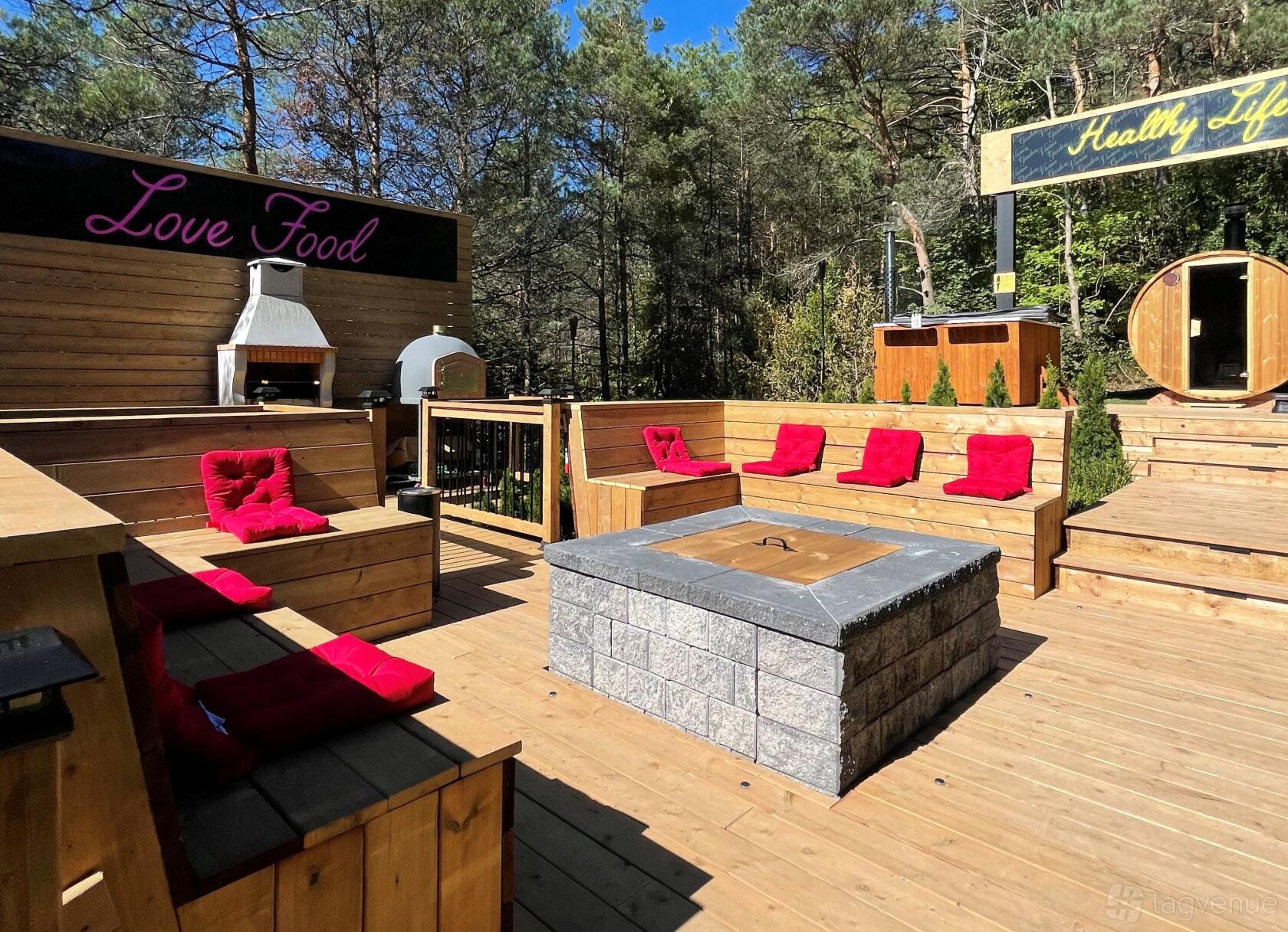 An outdoor event venue with wooden benches, red cushions, a central fire pit, and surrounding forest at Vivian Events & Spa.