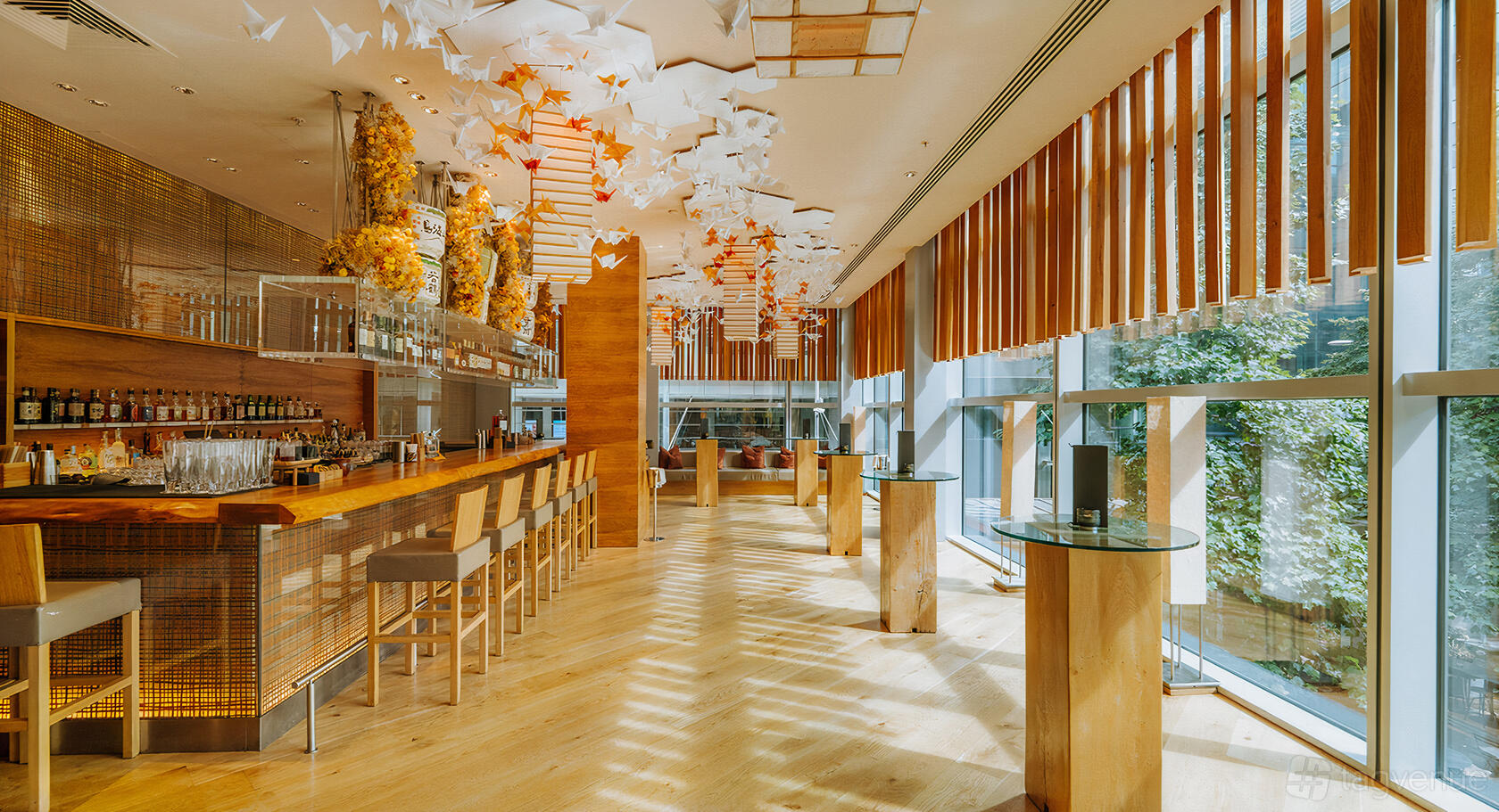 A lounge with floor-to-ceiling windows, high stools at a wooden bar, and hanging paper cranes at Roka Canary Wharf.