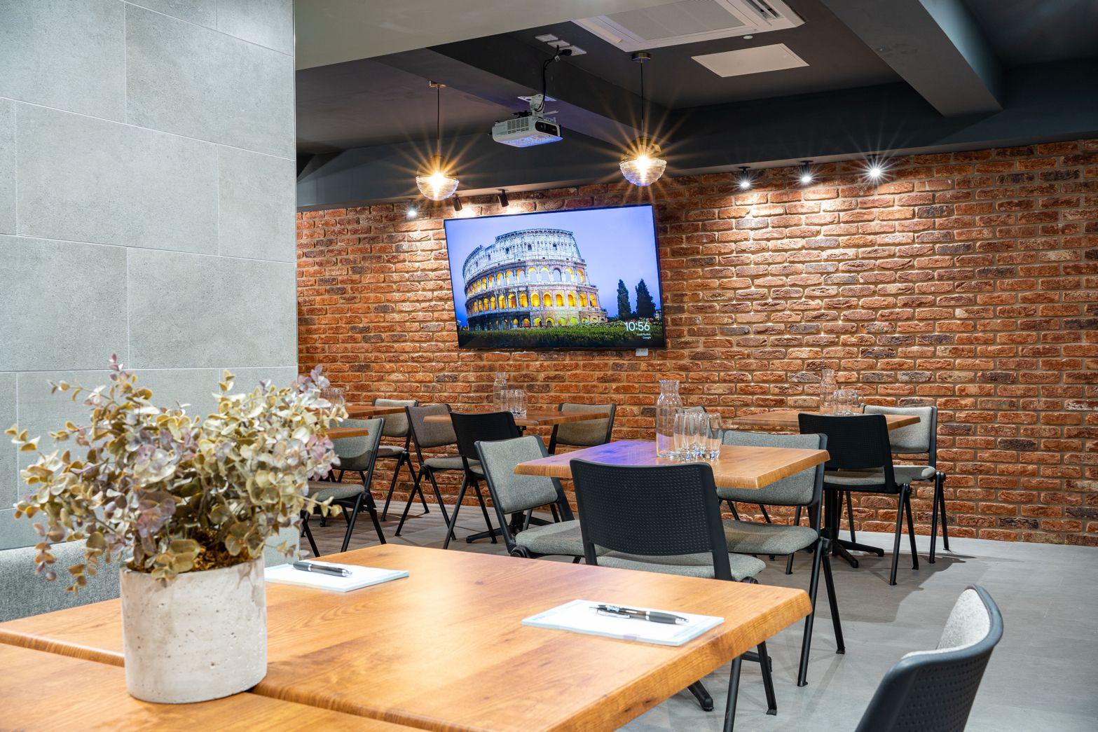 An event space with exposed brick walls, a mounted flat-screen, and wooden tables with black chairs at Wizu Workspace - Leeds - Park Place.