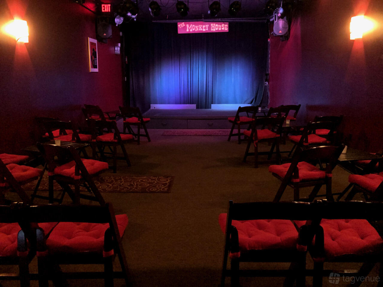A small theatre with cushioned folding chairs, red walls, and a stage with curtains at The Monkey House.