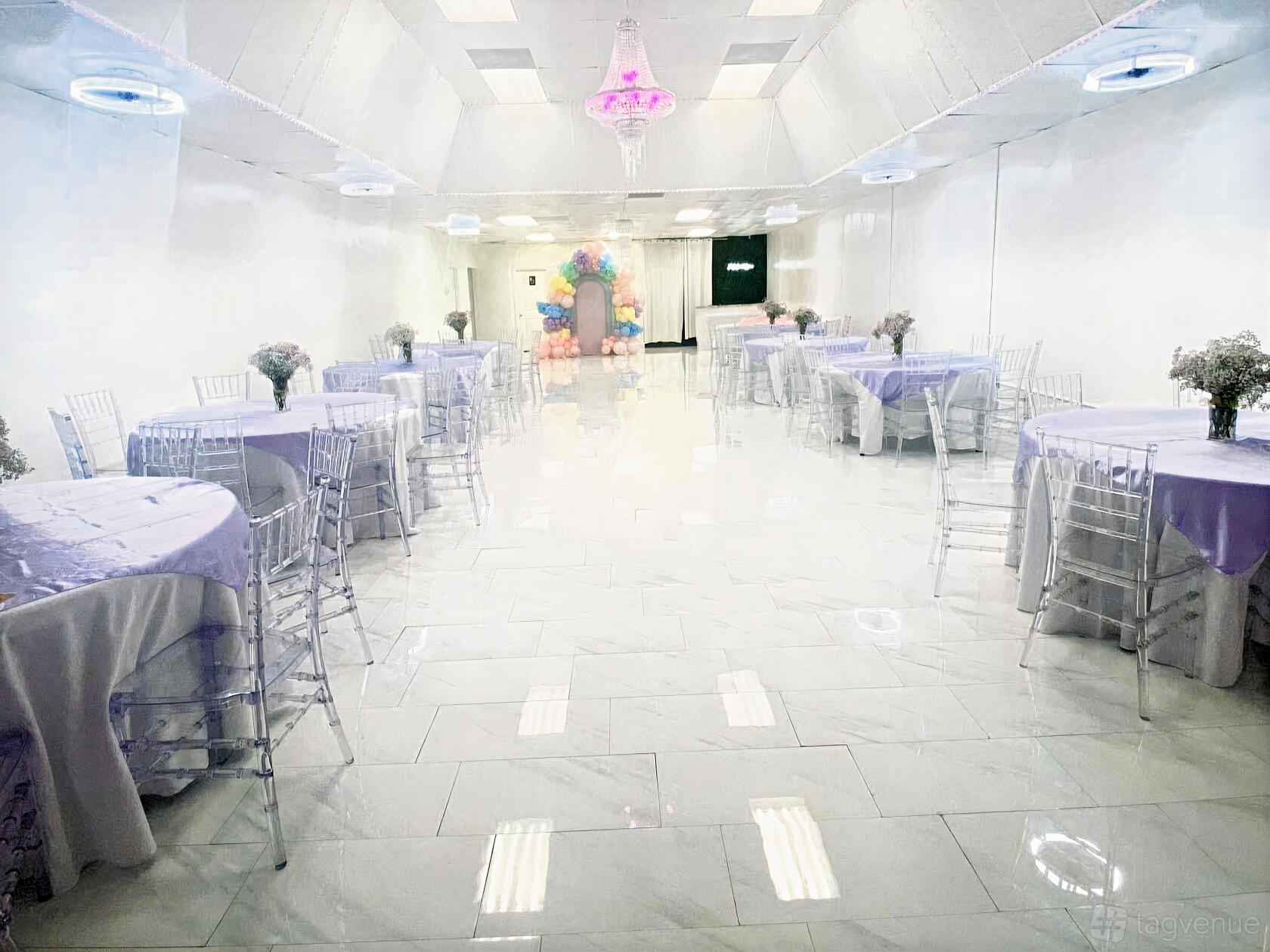 An event venue with lilac-covered round tables, clear chairs, and a pastel balloon arch at J&S Event Space.