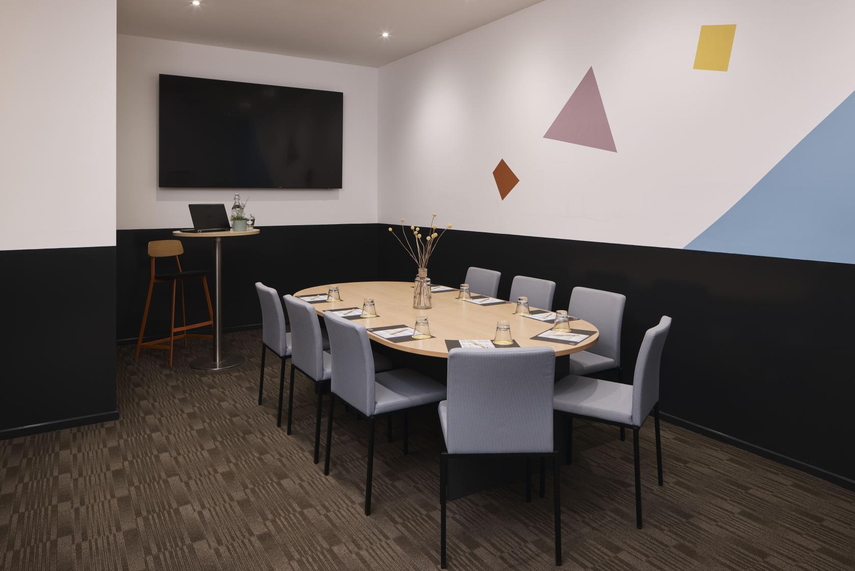 A hotel boardroom with an oval table, cushioned chairs, wall-mounted screen, and geometric wall art at Atura Dandenong.