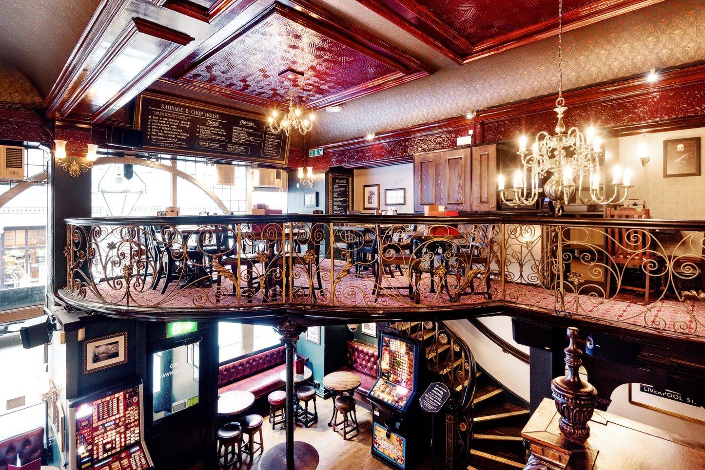 An event space in a pub with an ornate wrought iron mezzanine, red patterned ceiling, and chandelier at Lord Aberconway.