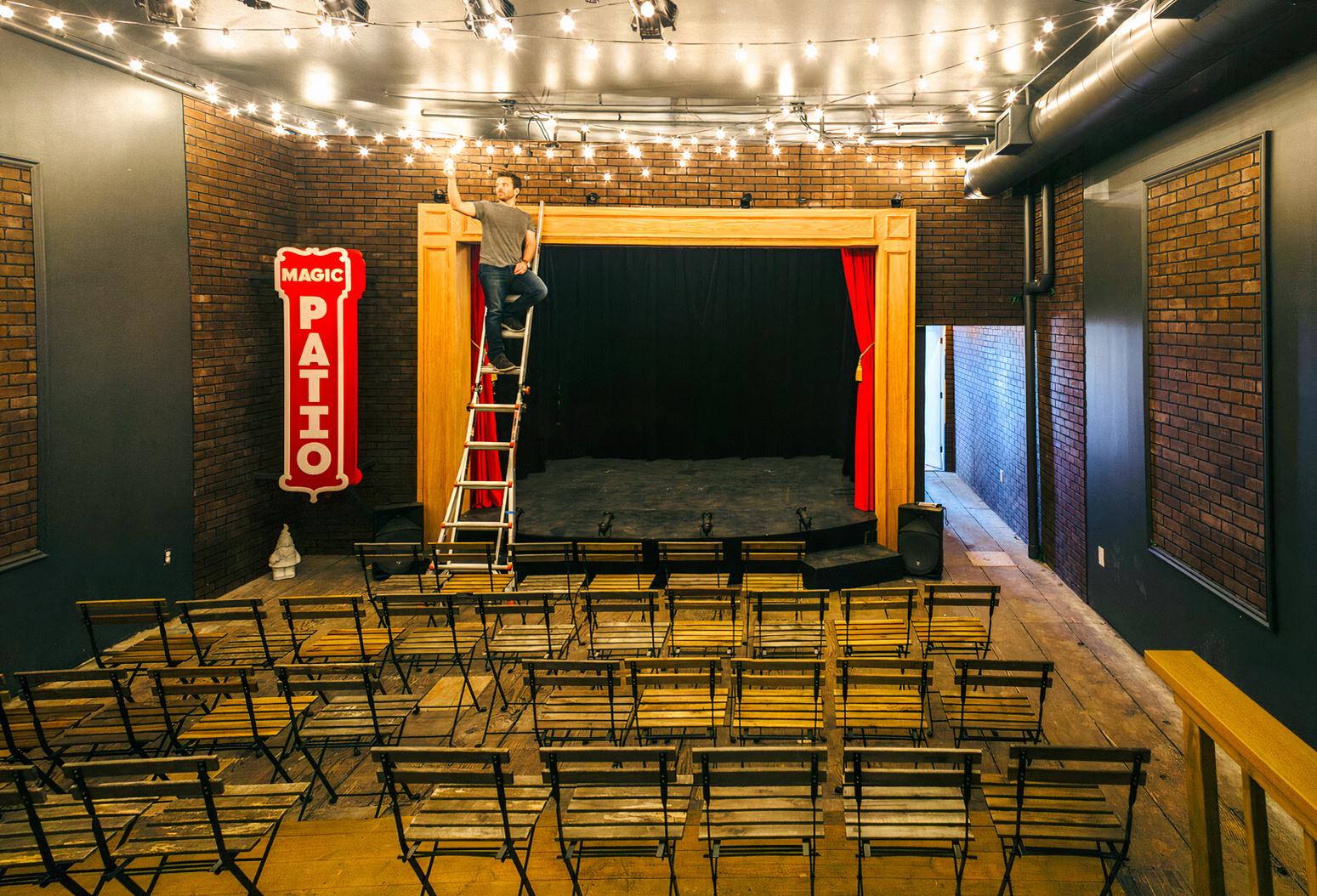 A theatre with rows of wooden chairs facing a stage with red curtains and exposed brick walls at The Magic Patio.
