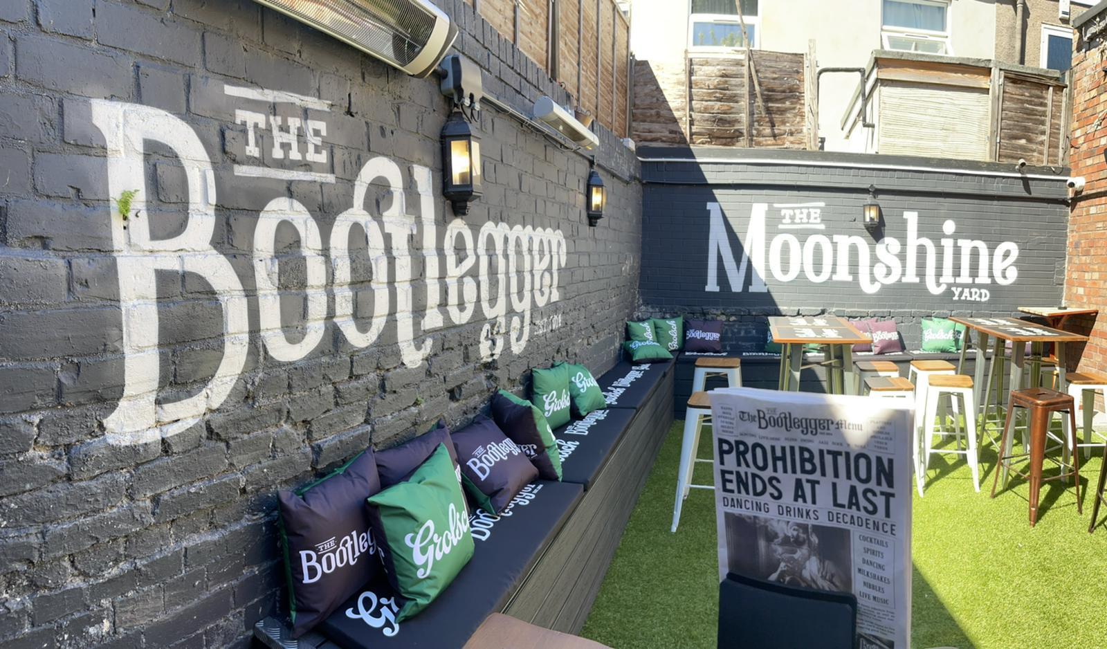 An outdoor bar area with artificial grass, wall seating, branded cushions, and painted signage at The Bootlegger.