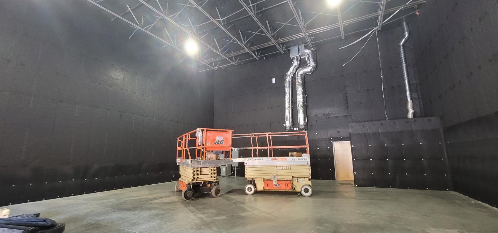 A film studio with high black acoustic walls, exposed ceiling pipes, and scissor lifts at Woman-owned black box stage and facility.