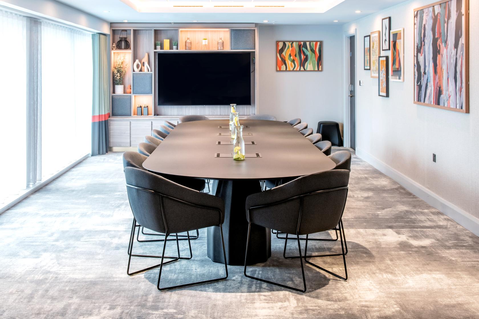 A boardroom with a long oval table, modern black chairs, large wall-mounted screen, and abstract art at Crowne Plaza Manchester City Centre.