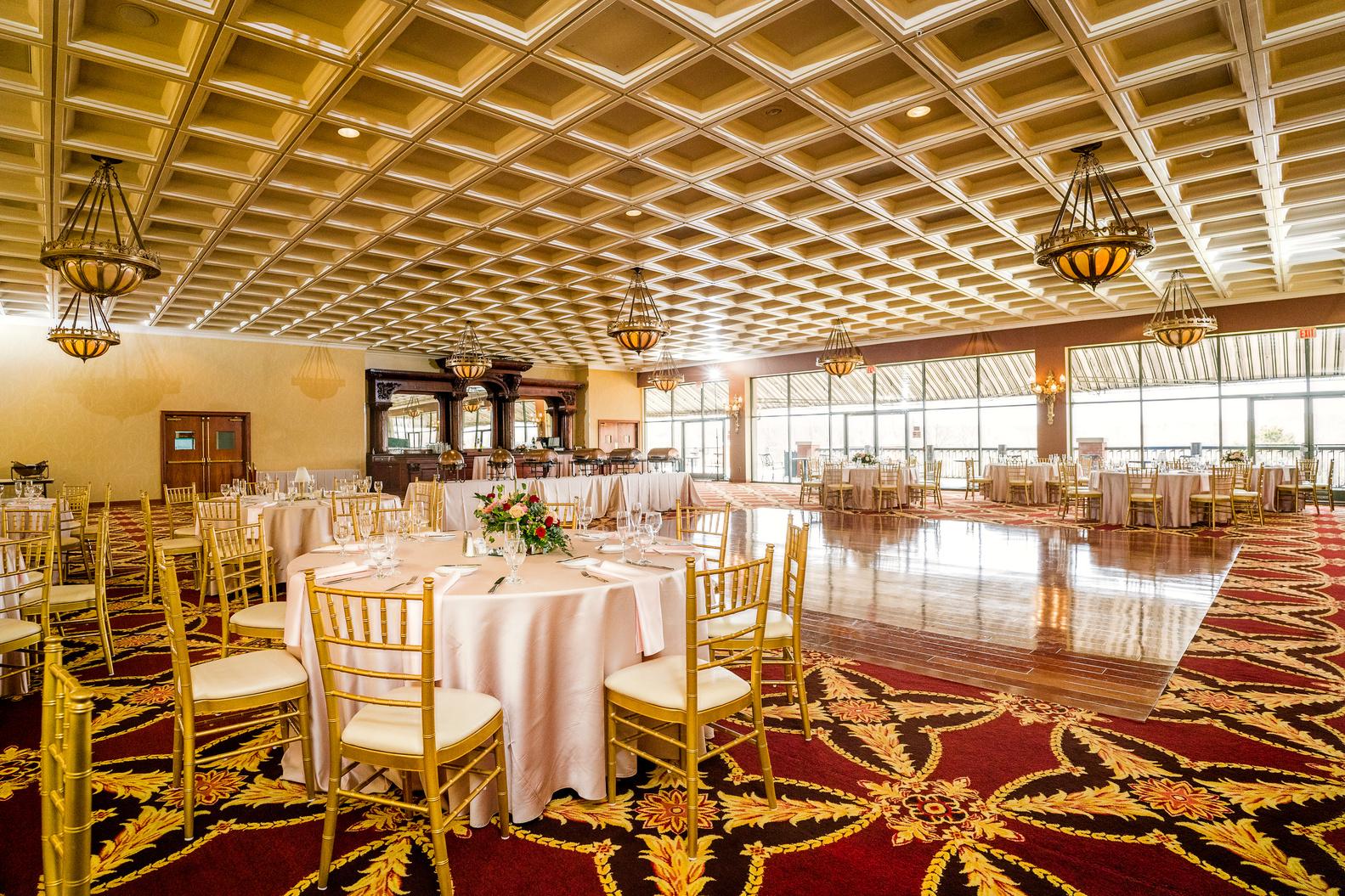 A ballroom with gold chandeliers, coffered ceiling, round tables with white linens, and patterned carpet at White Clay Creek Country Club.