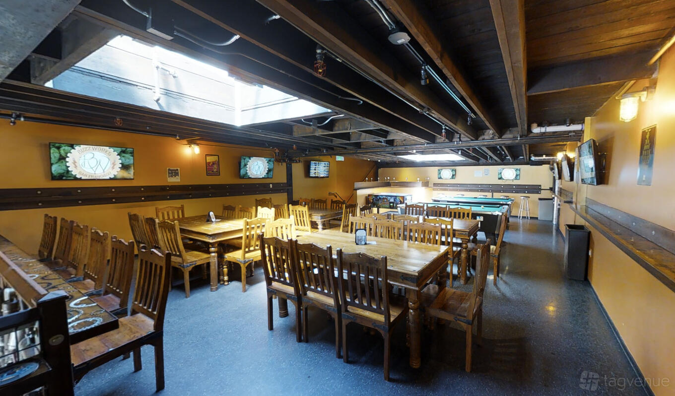 A bar with exposed wooden beams, natural light from a skylight, wooden tables, and wall-mounted TVs at BuzzWorks.