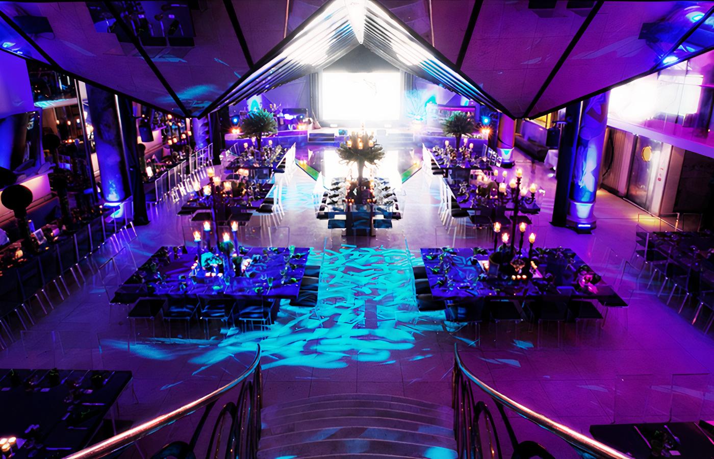A restaurant with dramatic colored lighting, long banquet tables, and a grand staircase at Quaglino's.