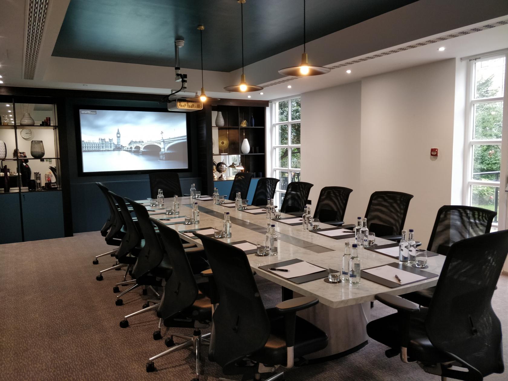 A hotel boardroom with a long marble table, ergonomic chairs, notepads, and a large screen at DoubleTree by Hilton London Kensington.