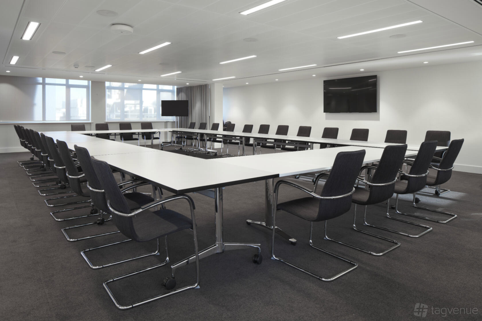 An auditorium with U-shaped tables, black chairs, wall-mounted screen, and large windows at 18 Smith Square.