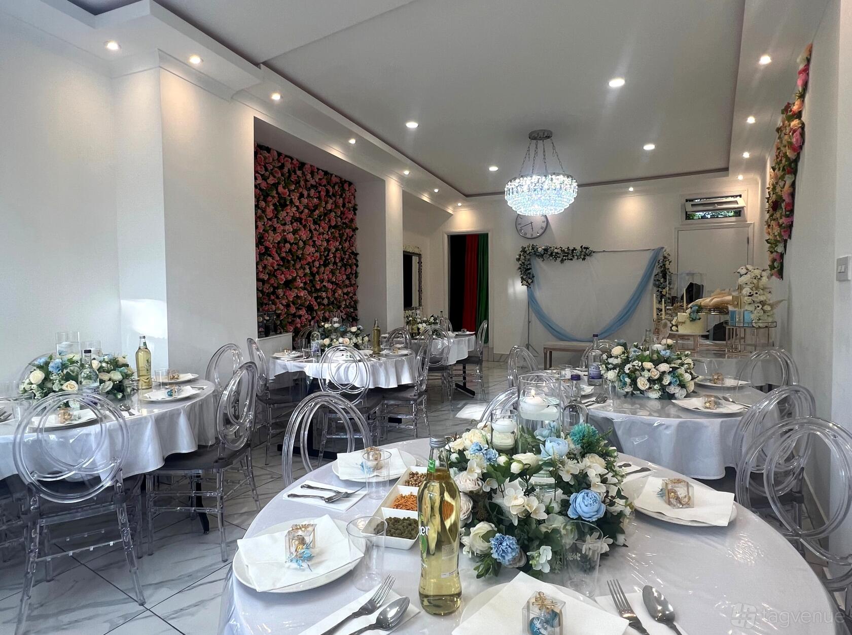 A hall with round tables, transparent chairs, floral centerpieces, and a crystal chandelier at Watandar Hall.