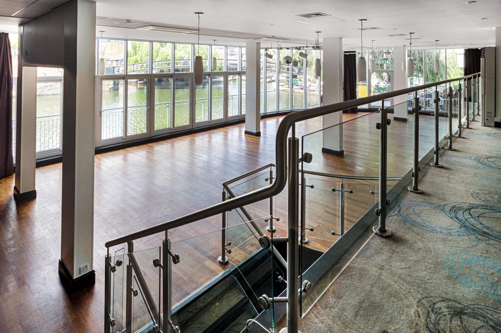A hotel event space with floor-to-ceiling windows, wooden flooring, and a glass railing at Glasshouse Camden.