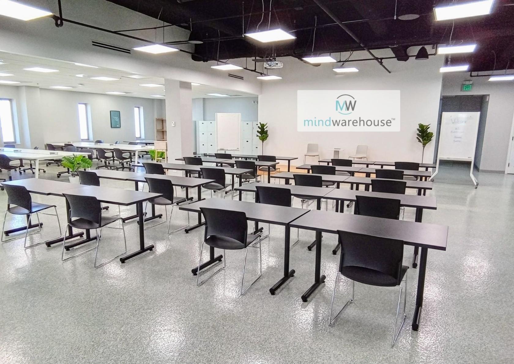 An office space with rows of black chairs and desks, potted plants, and large windows at mindwarehouse.