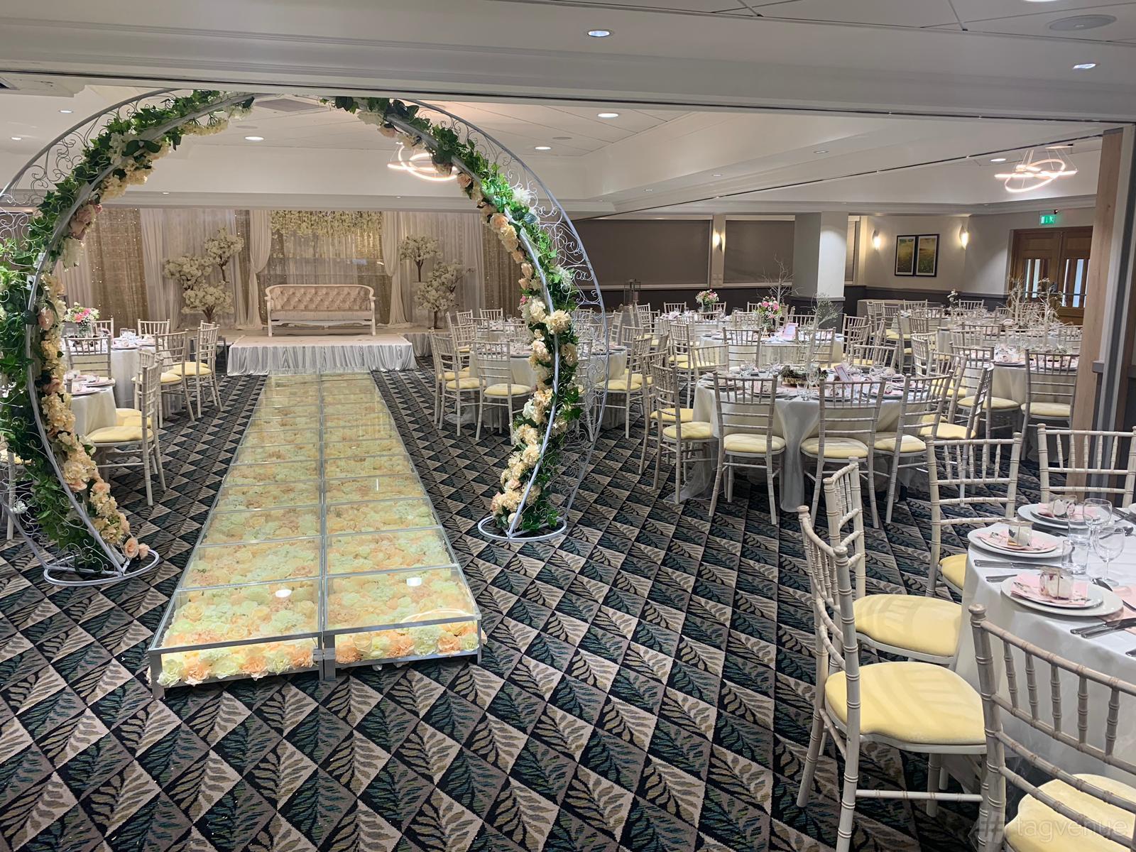 An event space with a floral archway, patterned carpet, round tables, and a raised stage at Holiday Inn Leicester - Wigston.
