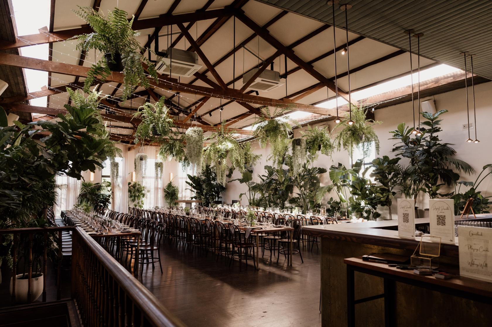 An event venue with abundant hanging greenery, exposed beam ceiling, and banquet tables at Glasshaus Brücke.