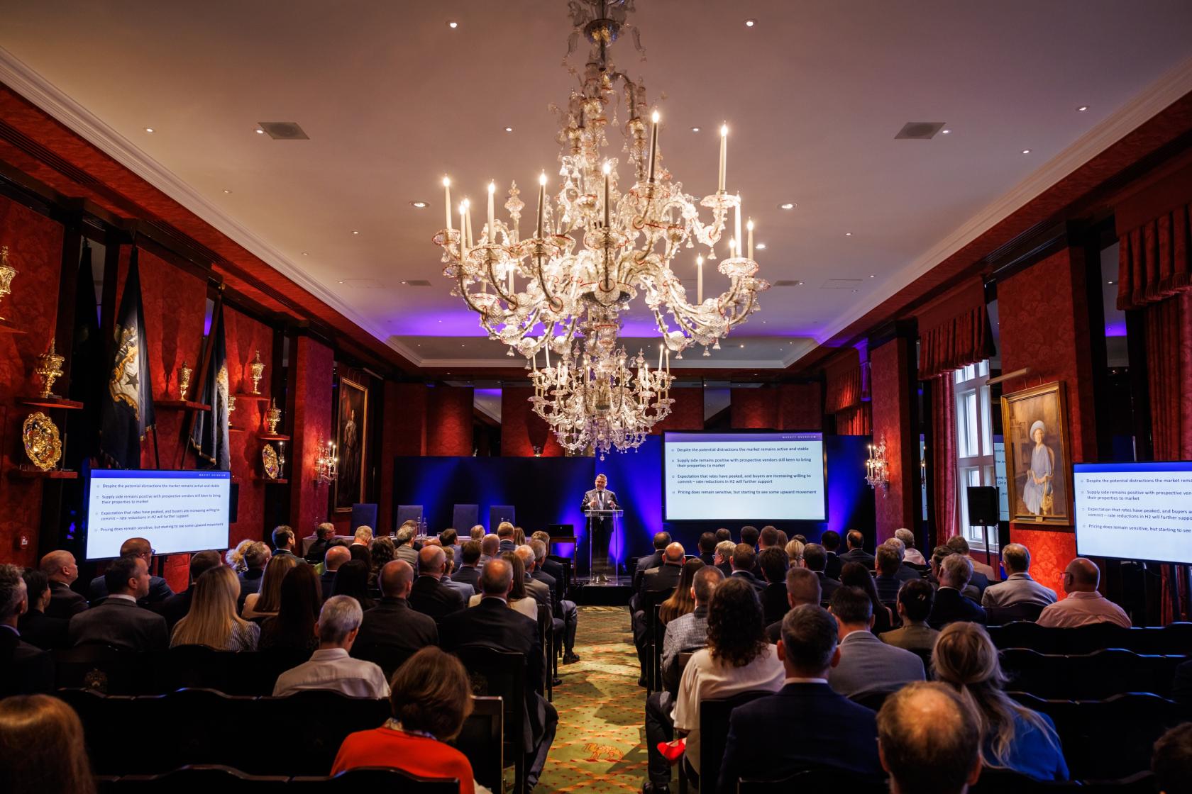 A hall with ornate chandeliers, red walls, and rows of chairs facing presentation screens at Grocers' Hall.