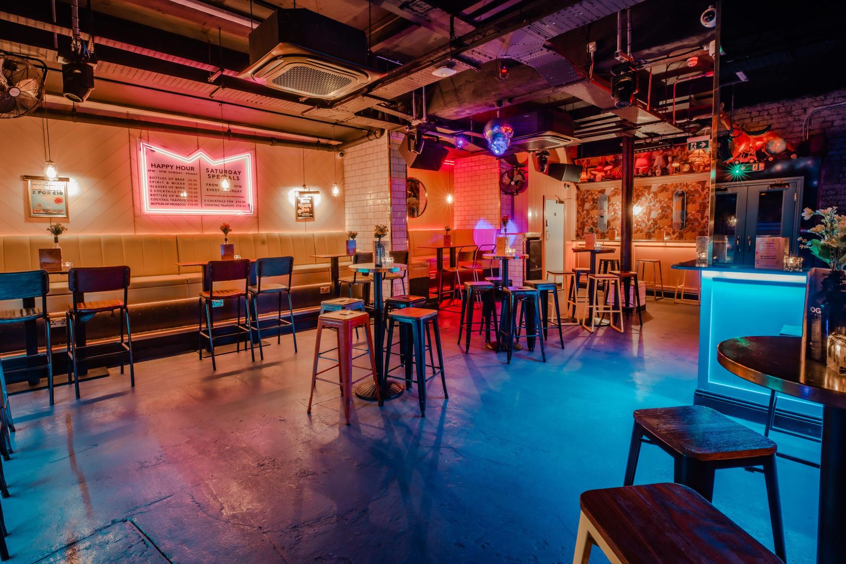 A cocktail bar with neon signs, high tables, bar stools, and colorful lighting at Simmons | Monument.