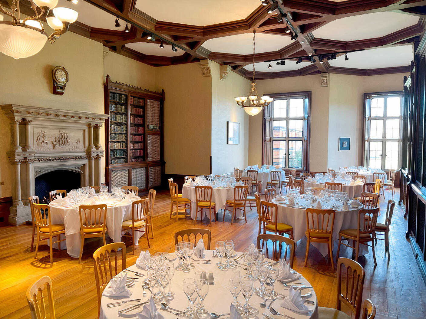 A hotel event room with tall sash windows, wood paneling, and round tables set with glassware in The Library at The Elvetham Hotel.