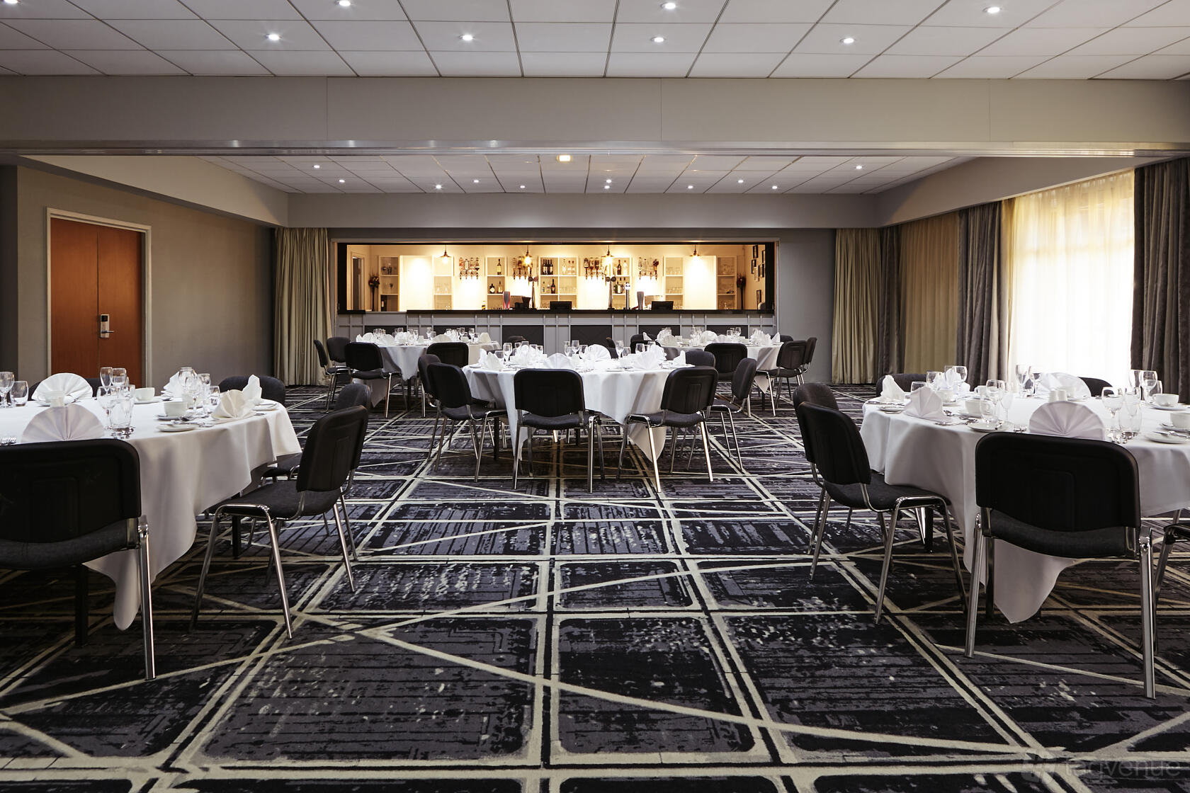 An event space with round tables dressed in white linens, patterned carpet, and a built-in bar at Novotel Manchester West.