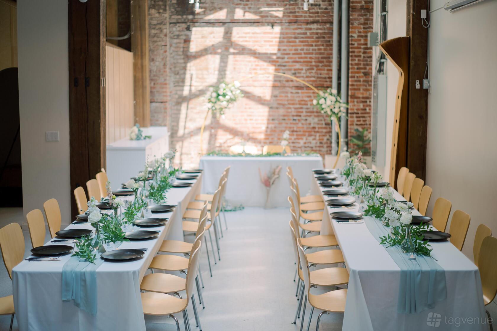 An event space with long tables draped in white linens, natural light, and exposed brick walls at This World's Ours Centre.