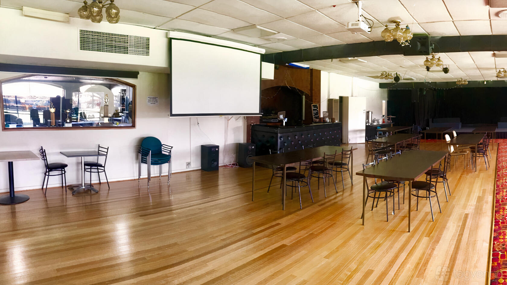 An event venue with polished wood floors, rectangular tables, a projector screen, and a mirrored bar at Flemington and Kensington Bowling Club.