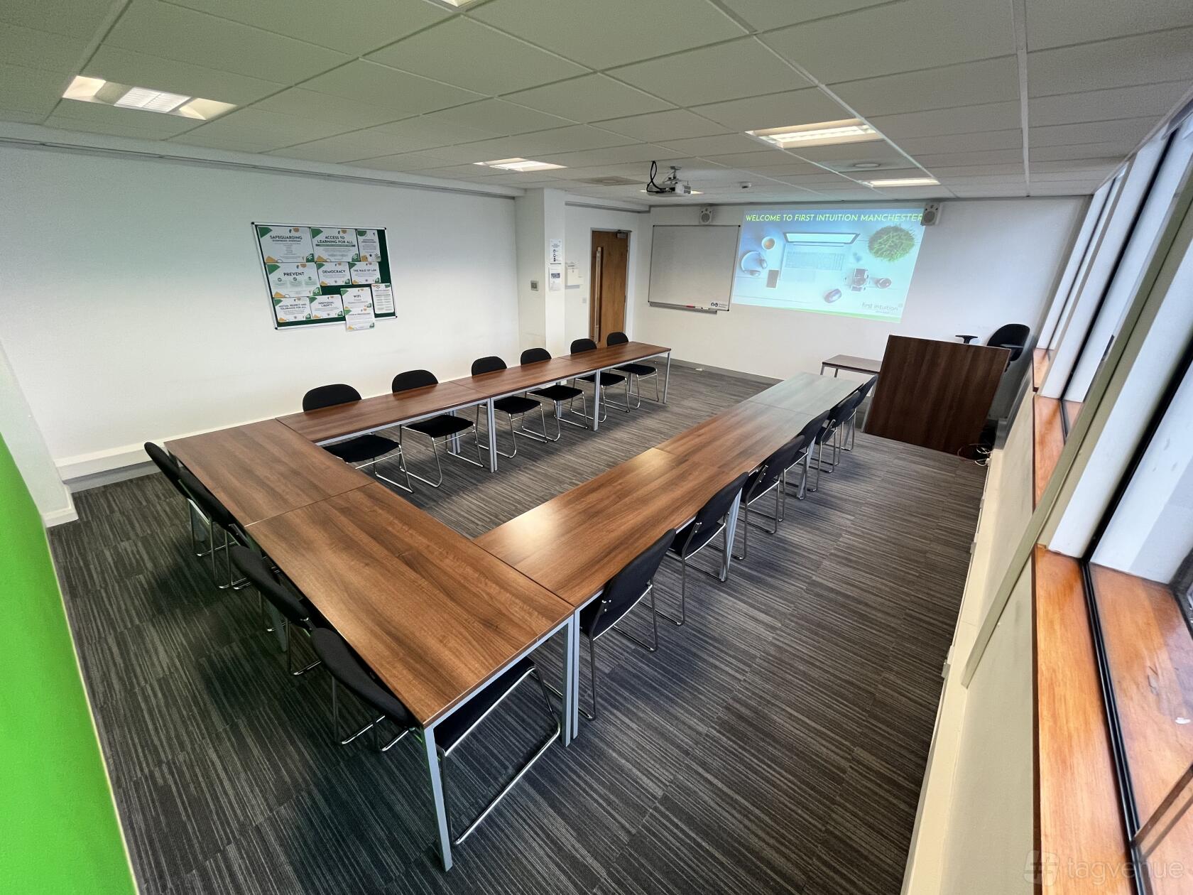 A meeting room with U-shaped wood tables, black chairs, a projector screen, and large windows at First Intuition Ltd Manchester.