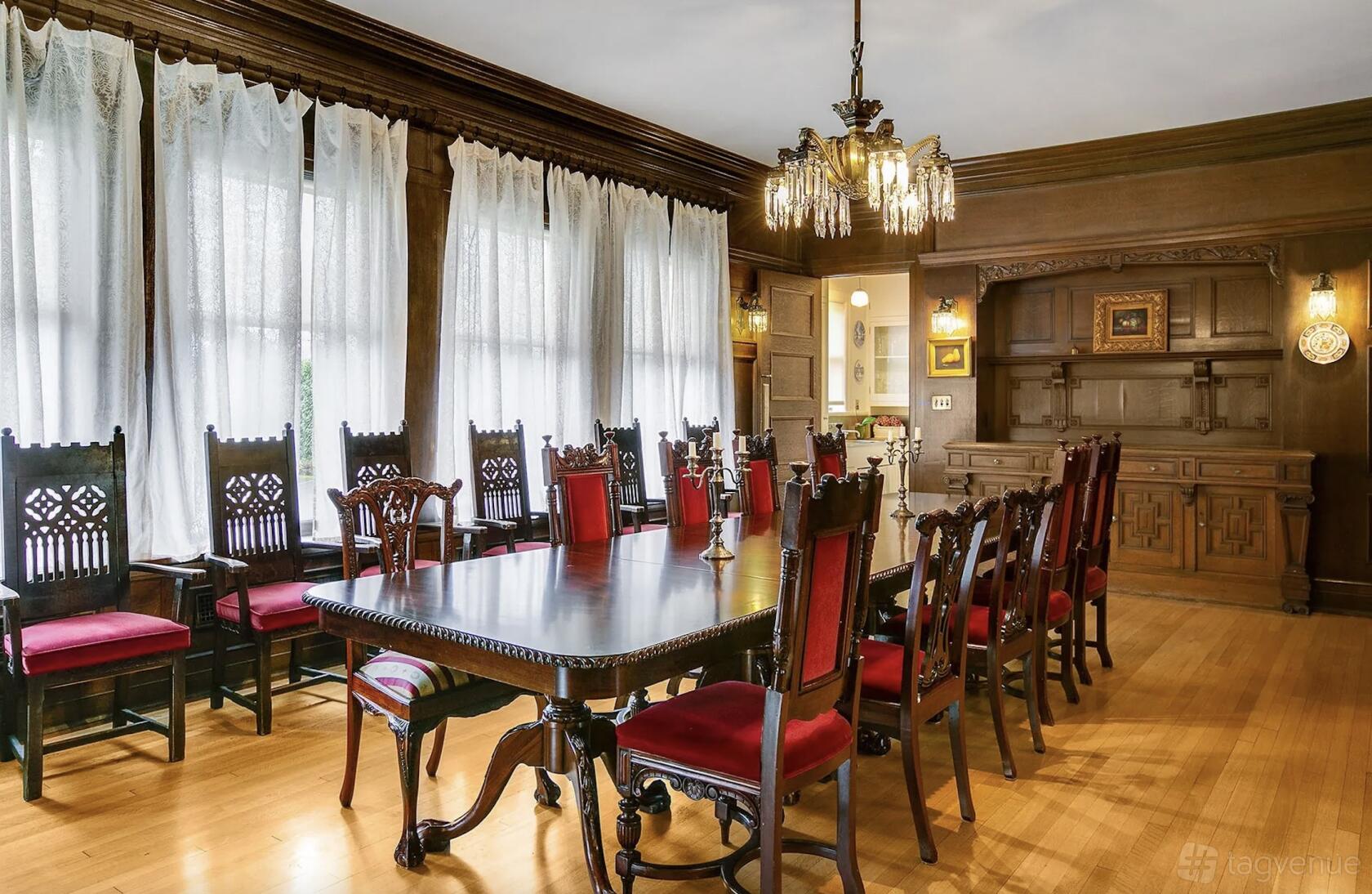 A house dining room with wood-paneled walls, a long table with red upholstered chairs, and chandeliers at Shafer Baillie Mansion Bed & Breakfast.
