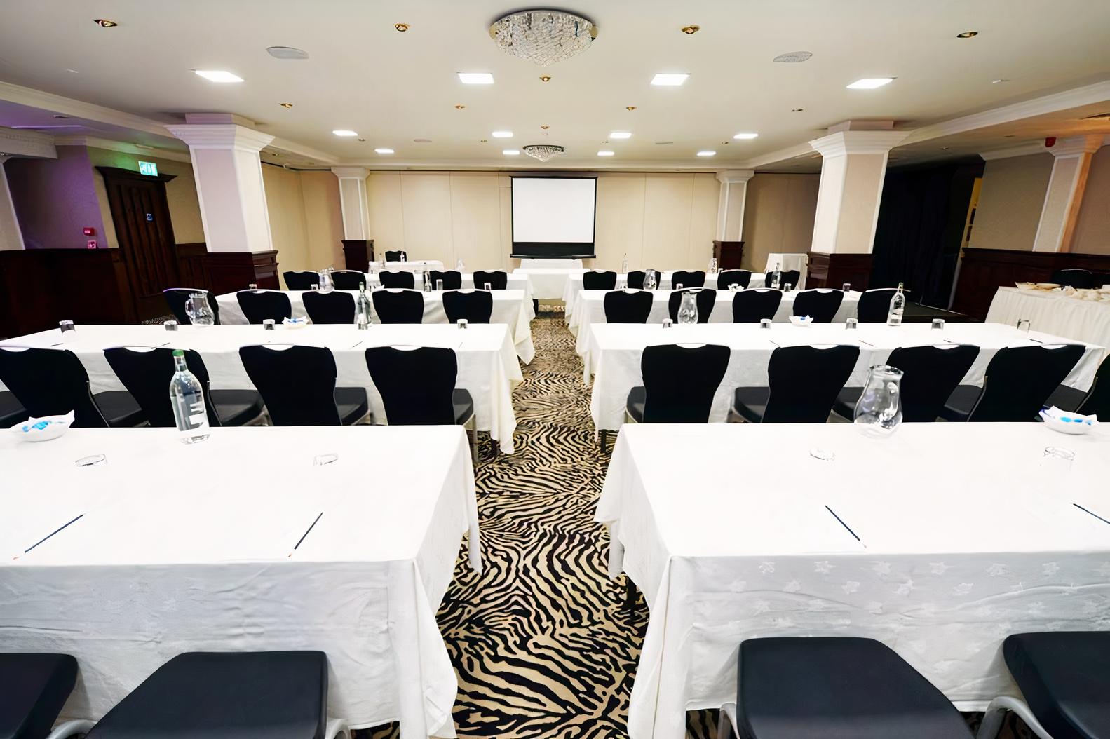 An event space with zebra-patterned carpet, white-covered tables in classroom setup, and a projector screen at The Glynhill Hotel.
