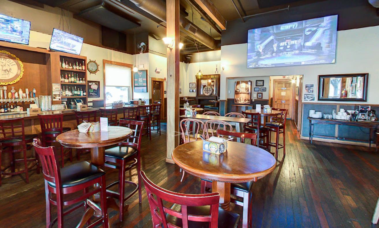 A bar with wooden tables, high-top seating, mounted TVs, and exposed ceiling pipes at Seawolf Public House.