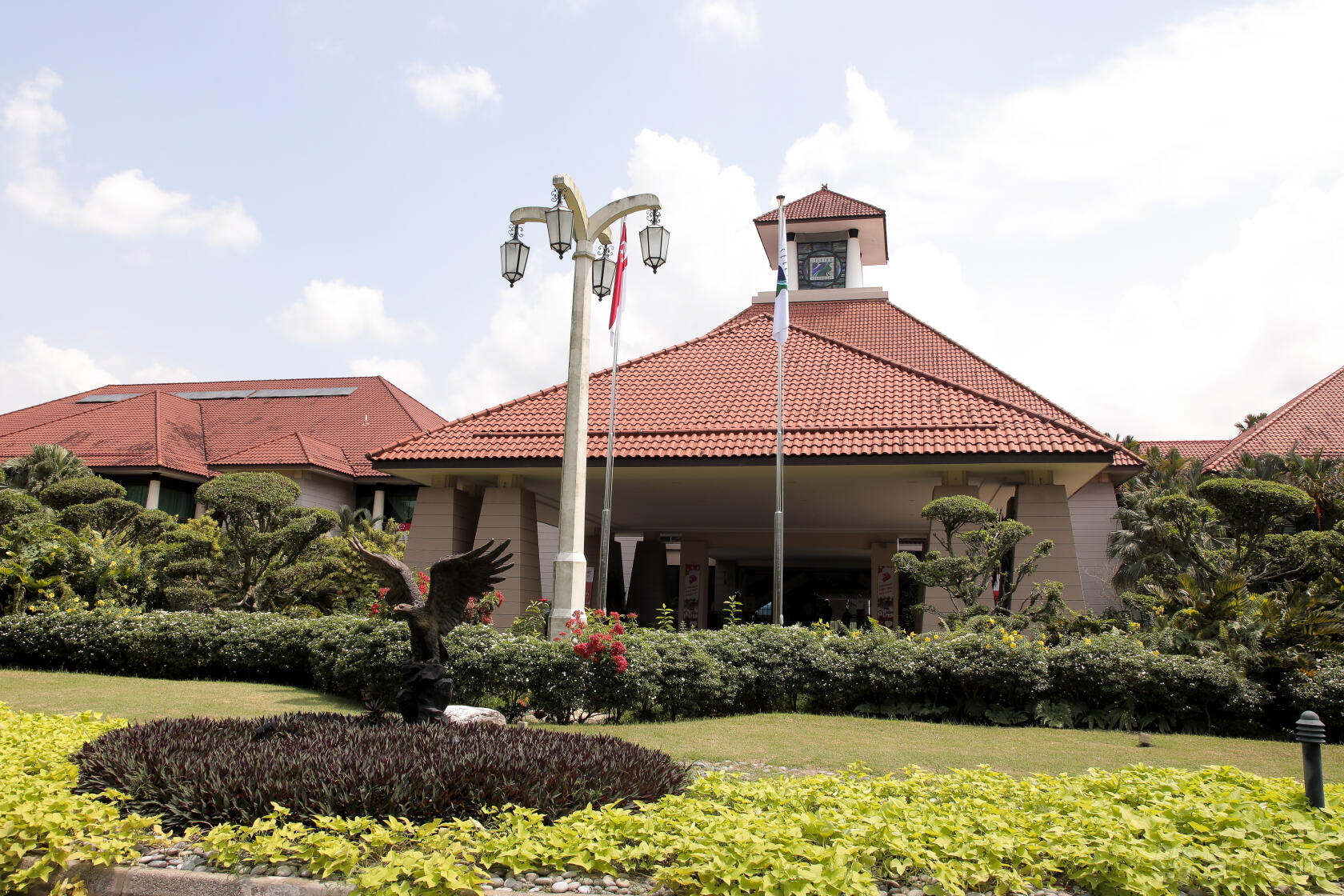 A golf club entrance with manicured hedges, a clock tower, red-tiled roof, and flagpoles at Seletar Country Club.