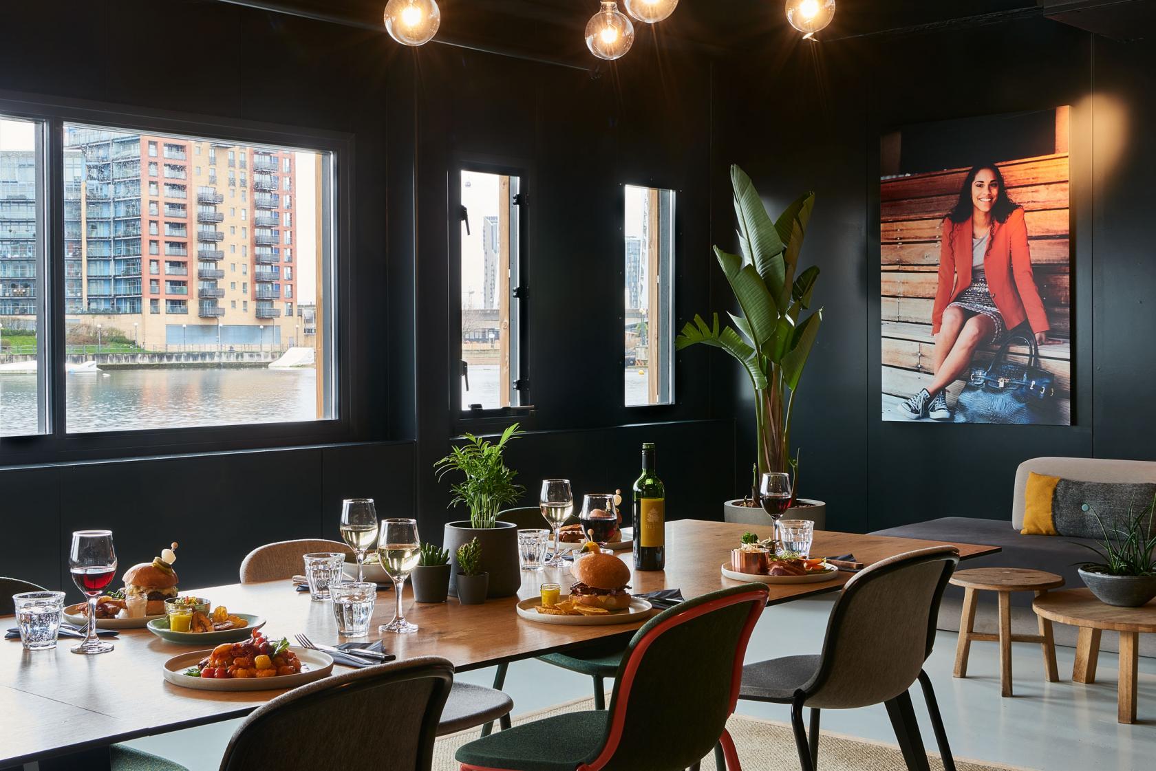 A hotel meeting space with large windows overlooking the water, wooden table set for dining, and modern decor at Good Hotel London.
