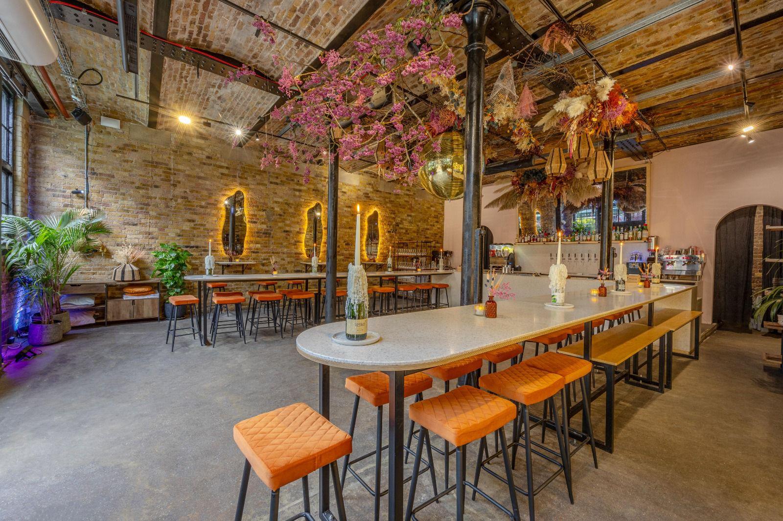 An event space in a bar with exposed brick walls, floral ceiling decor, and long tables with orange stools at Ester.