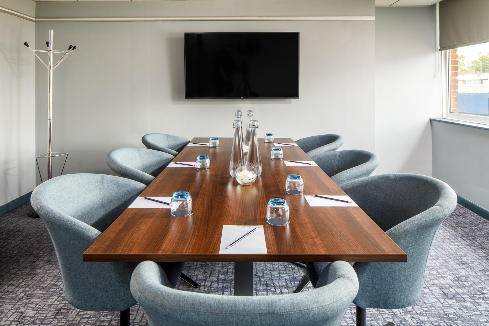 A meeting room with a wooden conference table, upholstered chairs, and a wall-mounted screen at Harlow Hotel by Accor.